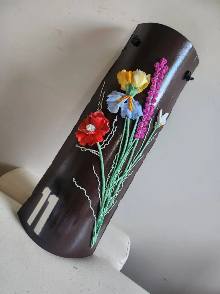 Collezioni Una targa marrone scuro con il numero 11 in bianco e motivi floreali colorati in rilievo, tra cui fiori rossi, gialli, blu, viola e bianchi, appoggiata a una parete chiara e a un divano.
