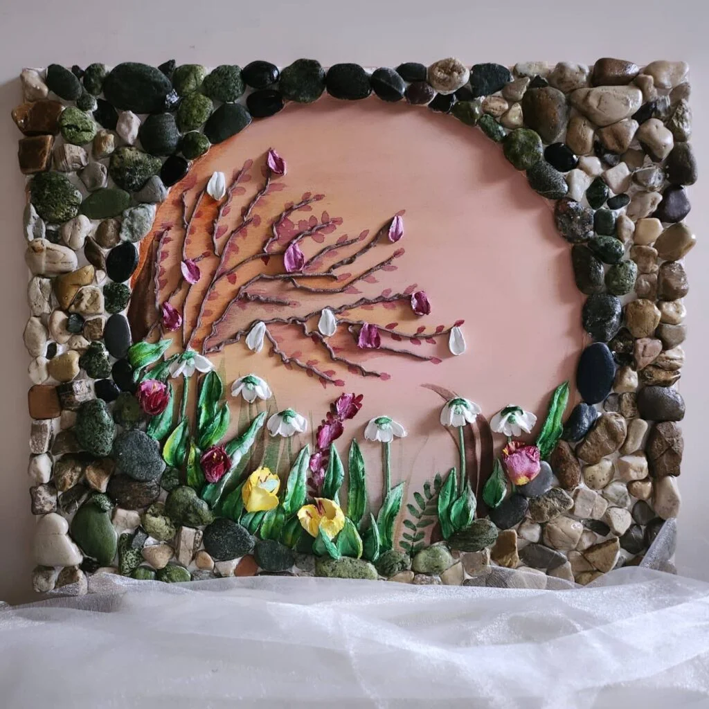 Una cornice decorativa realizzata con pietre di vario tipo circonda un'opera d'arte floreale di forma circolare, con fiori e foglie dipinti nei toni del rosa, del bianco e del giallo su uno sfondo color pesca.